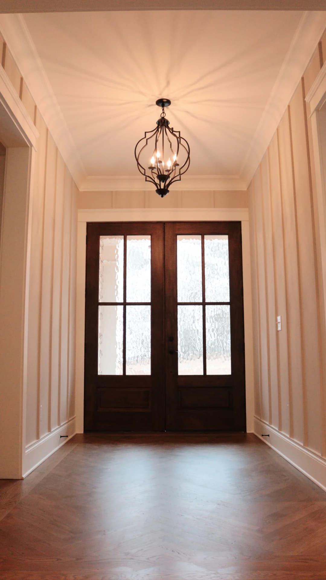 Wood and glass double front doors beneath a warm chandelier in a finished home